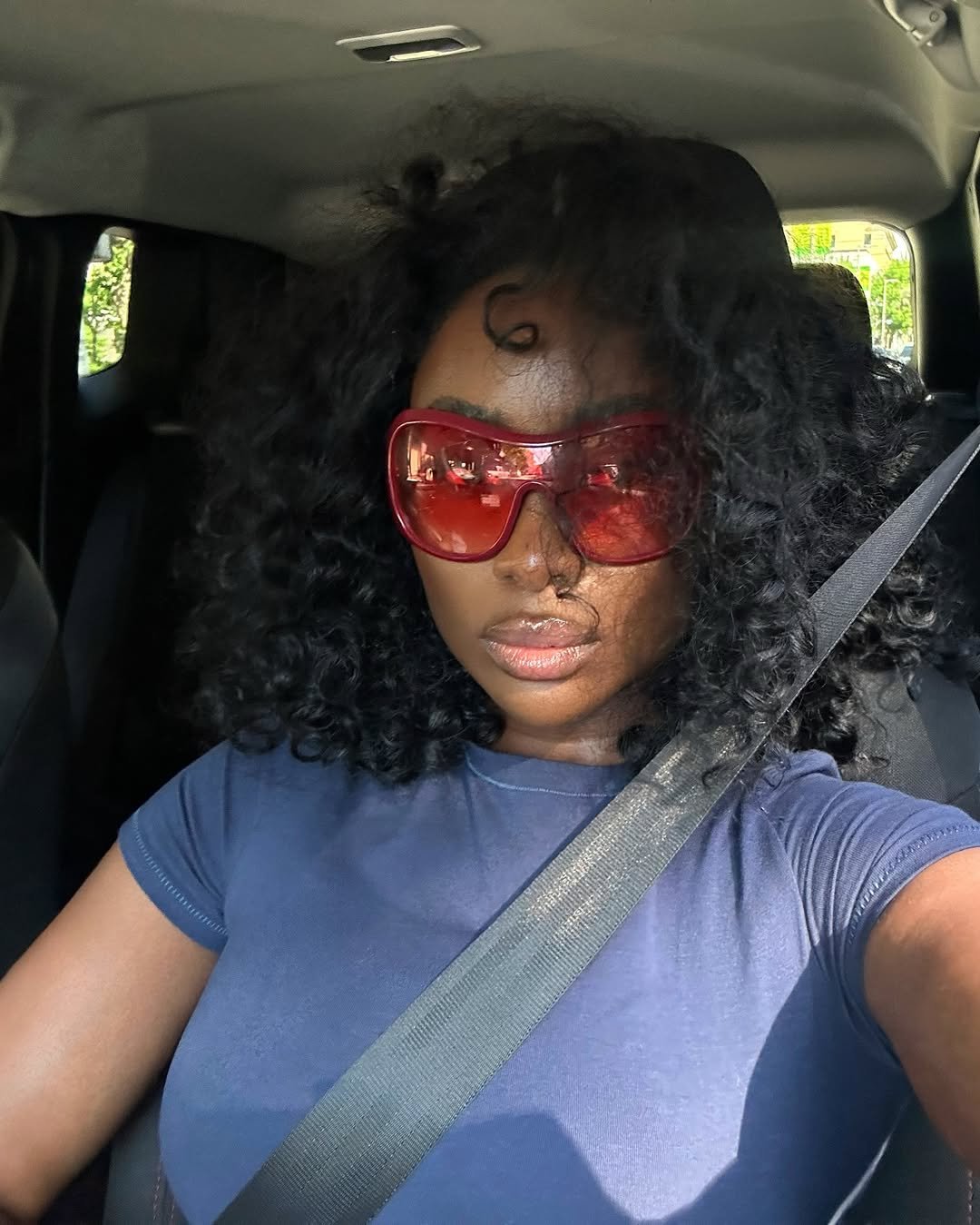 Mulher com cabelo cacheado usando óculos de sol do momento vermelhos, sentada dentro de um carro.