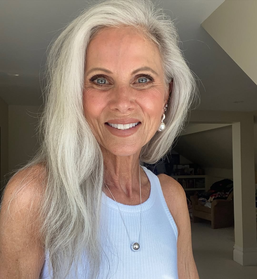 Mulher madura sorridente com cabelo grisalho e olhos marcantes, desafiando padrões convencionais de beleza em ambiente interno.