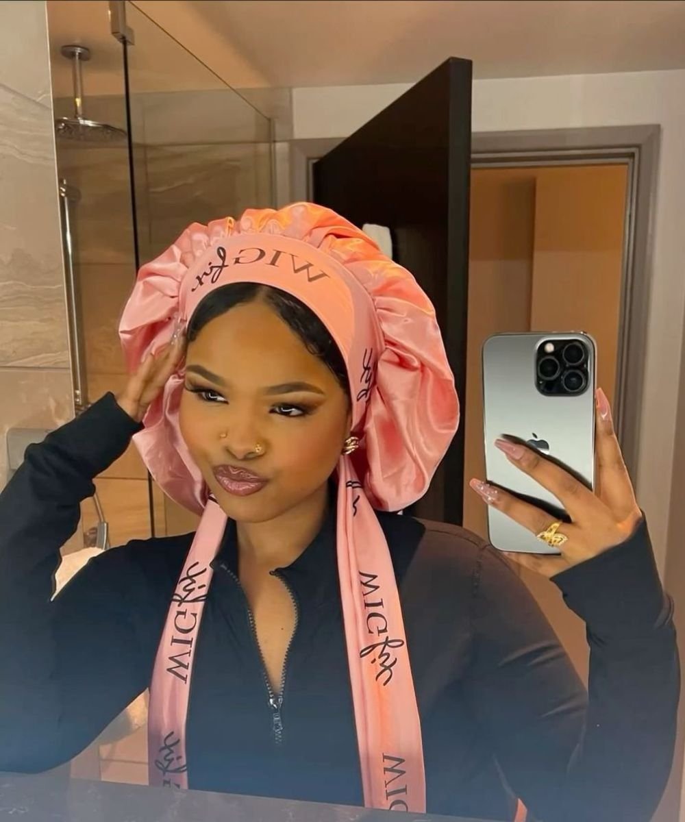 Usuária usando touca de cetim rosa, posando para selfie em banheiro, com smartphone na mão.