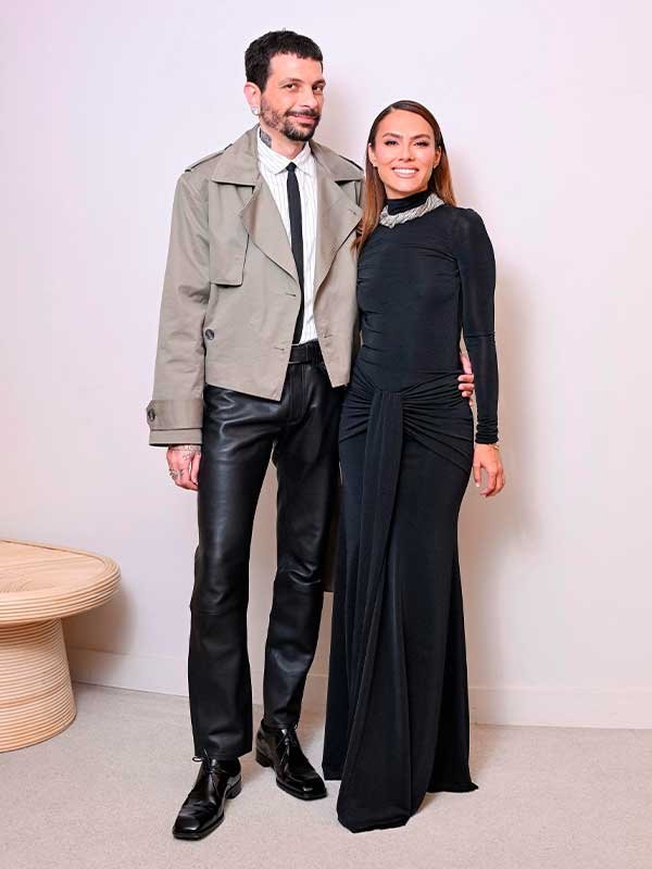 Um homem e uma mulher posam juntos. Ela veste um vestido preto elegante e ele usa uma jaqueta cinza. PatBO e Eduardo Caires.