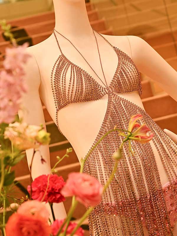 Vestido metálico com franjas, cercado por flores vibrantes; design sofisticado da PatBO.