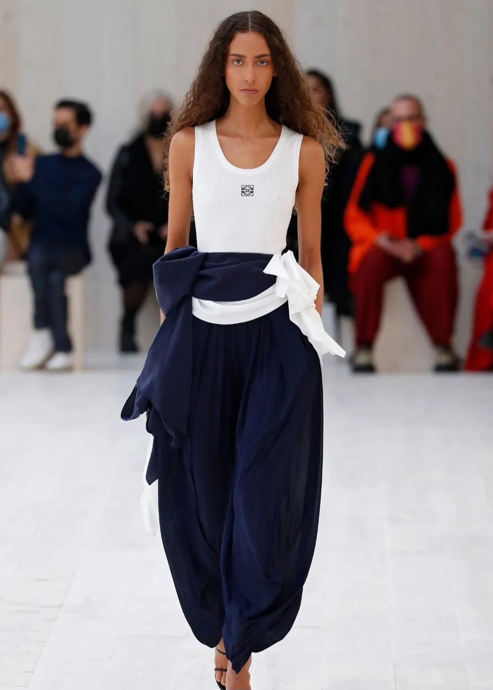 Desfile de moda com modelo usando regata branca e calça larga azul-marinho.