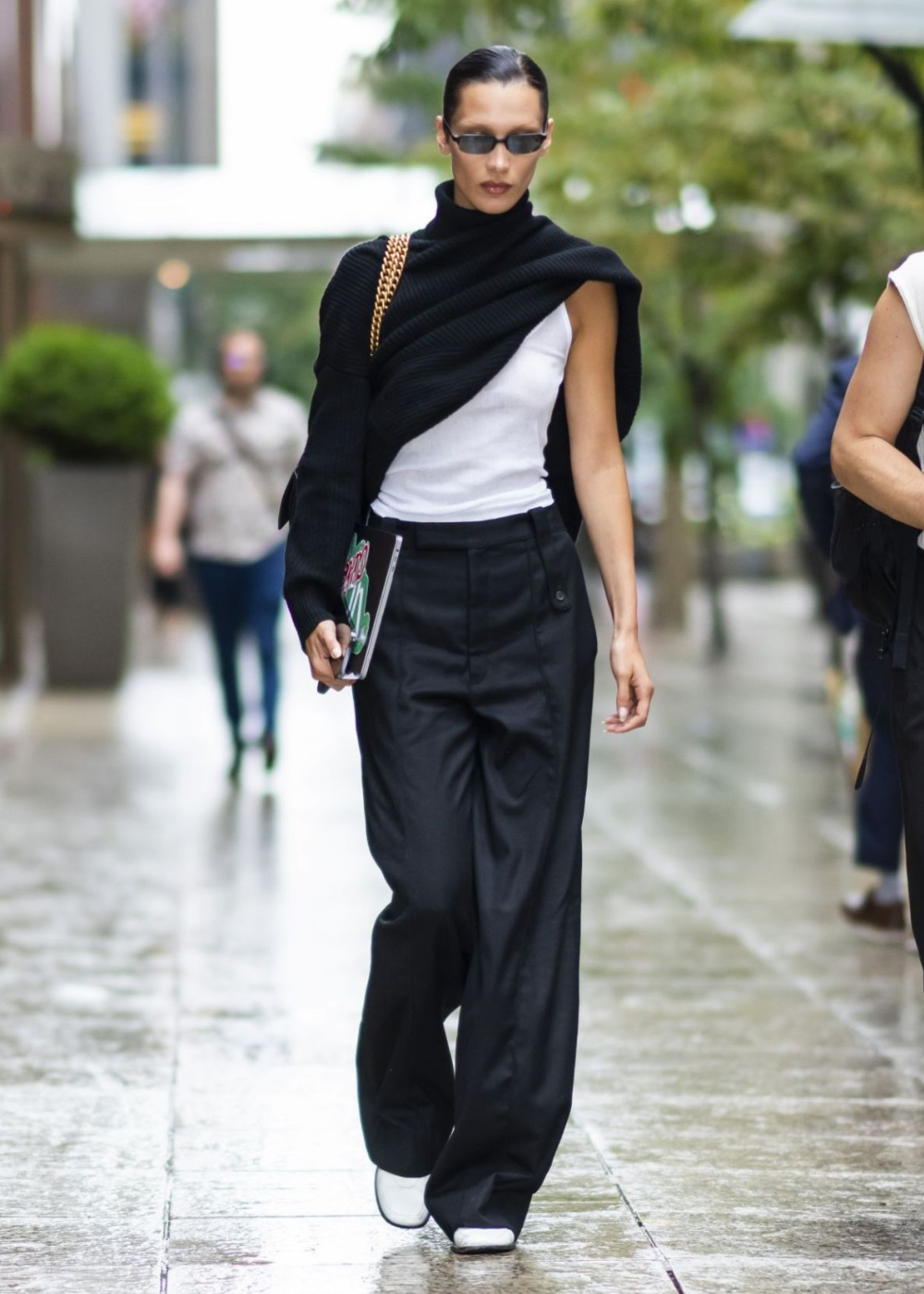 Pessoa usando regata branca e calças pretas largas, com suéter preto, caminhando na rua.