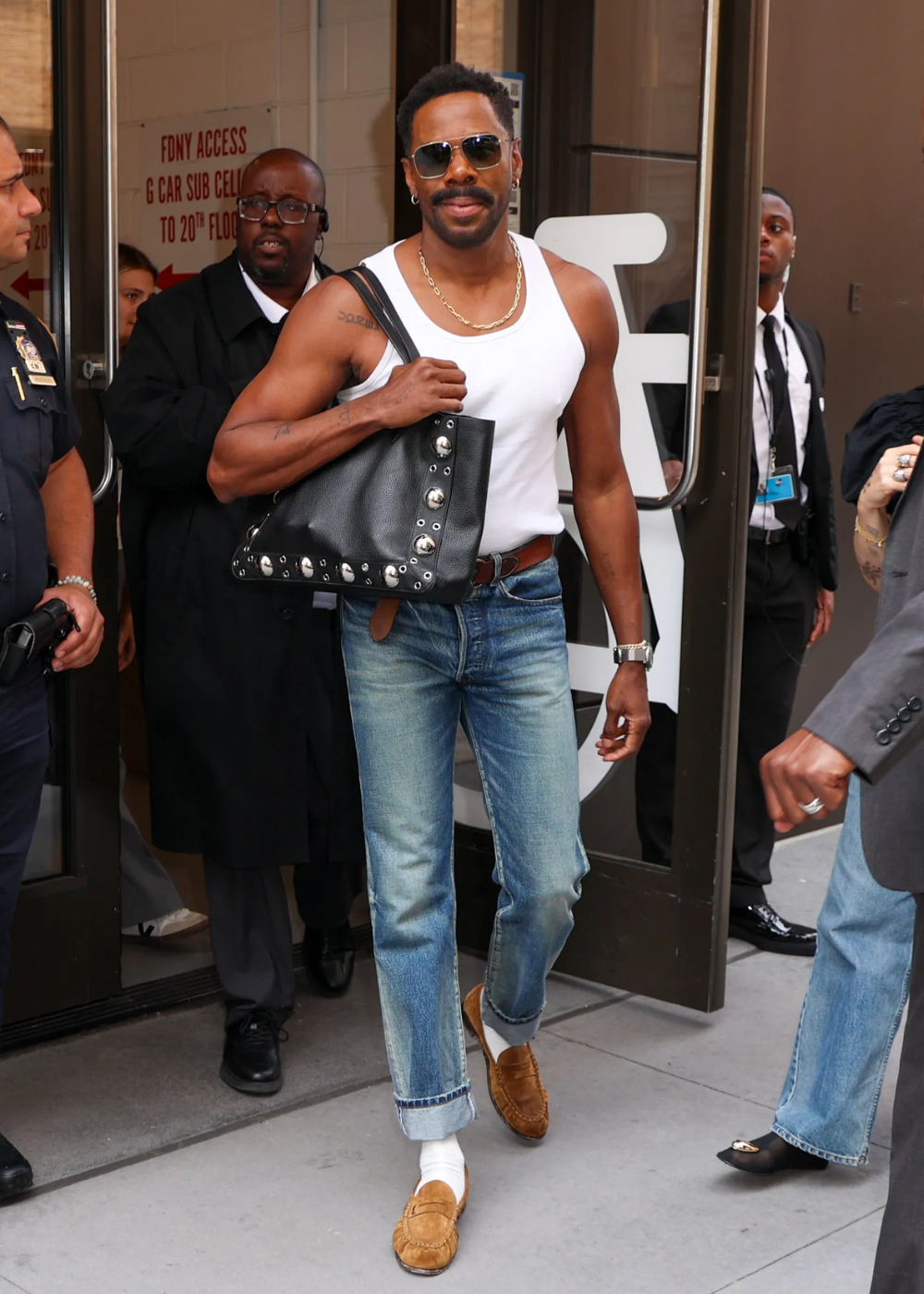 Homem de regata branca e jeans, carregando bolsa preta estilizada. Usa óculos escuros e mocassins marrons.