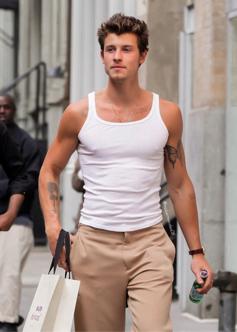 Homem com regata branca, calça bege, carregando sacola e garrafa em ambiente urbano.