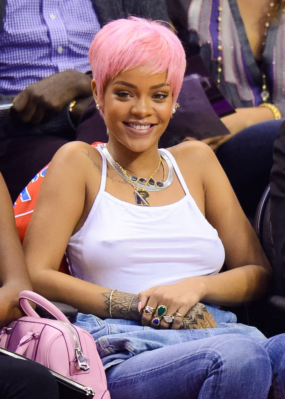 Pessoa sorrindo com cabelo rosa curto usando regata branca, colares e anéis, sentada em evento.