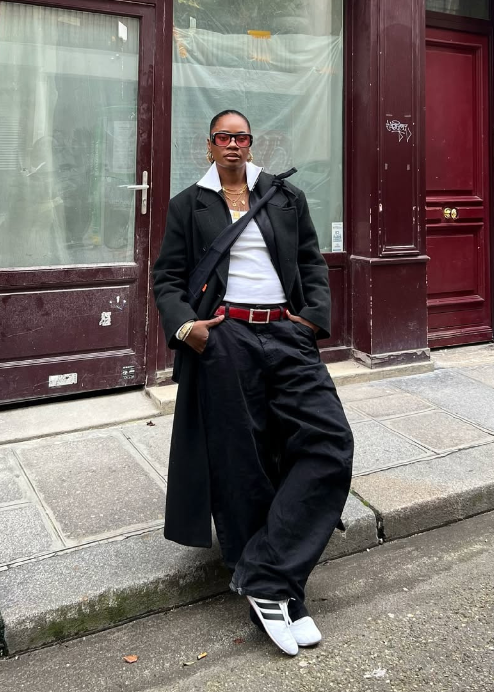 Pessoa com casaco preto, regata branca, calças largas, óculos escuros e sapatos brancos encostada em uma parede vermelha.