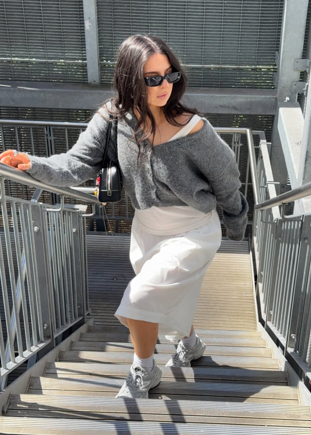 Mulher subindo escada usando suéter cinza e saia branca, segurando corrimão. Óculos escuros e cabelo castanho solto.