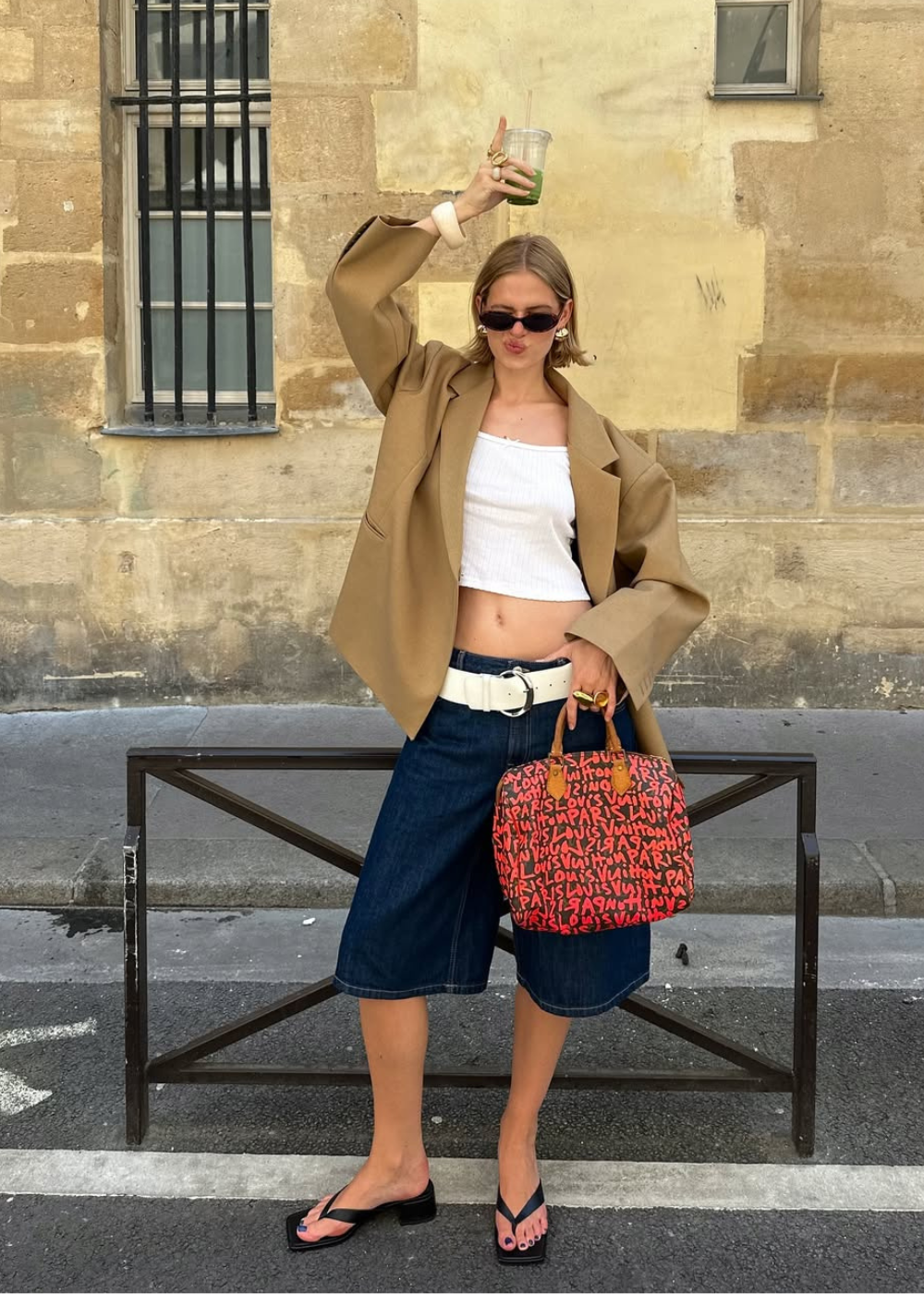 Pessoa elegante com regata branca, jaqueta bege e bermuda jeans. Carrega uma bolsa colorida e segura bebida.