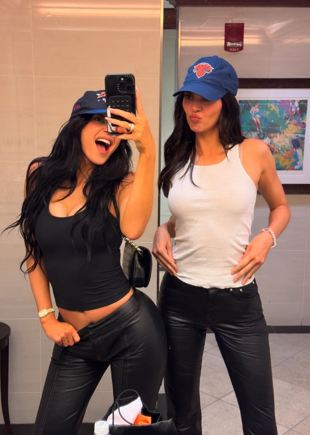 Mulher de regata branca e boné azul posa ao lado de amiga em regata preta, ambas sorrindo.