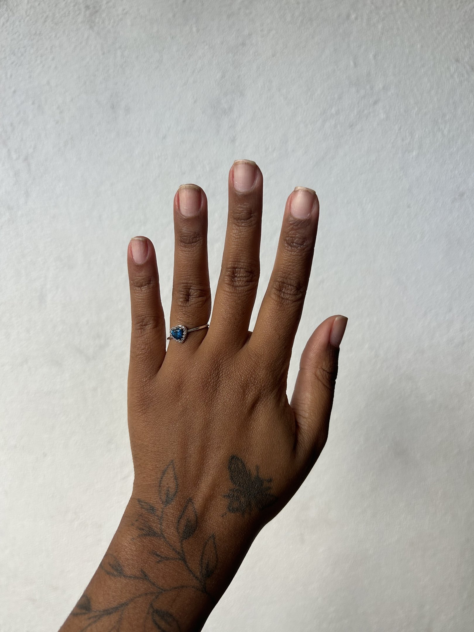 Mão com tatuagem, anel azul e unhas curtas. Não há unha autocolante visível.
