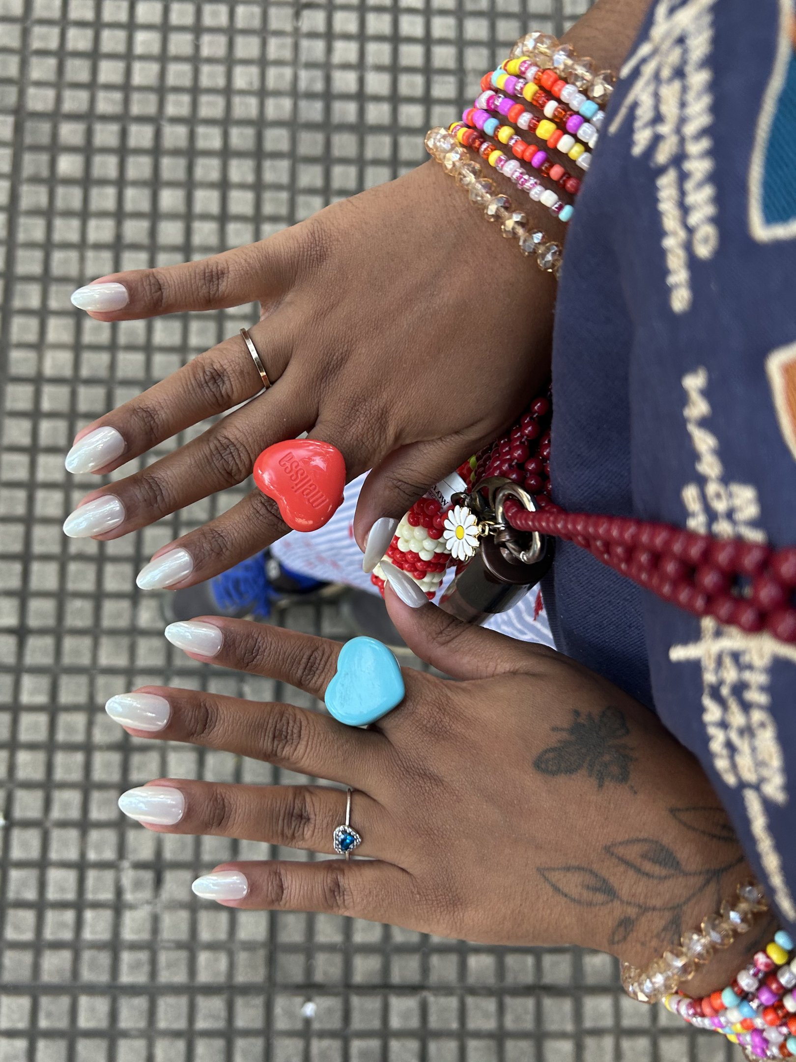 Mãos com unha autocolante e anéis coloridos. Braceletes de miçangas complementam o visual vibrante.