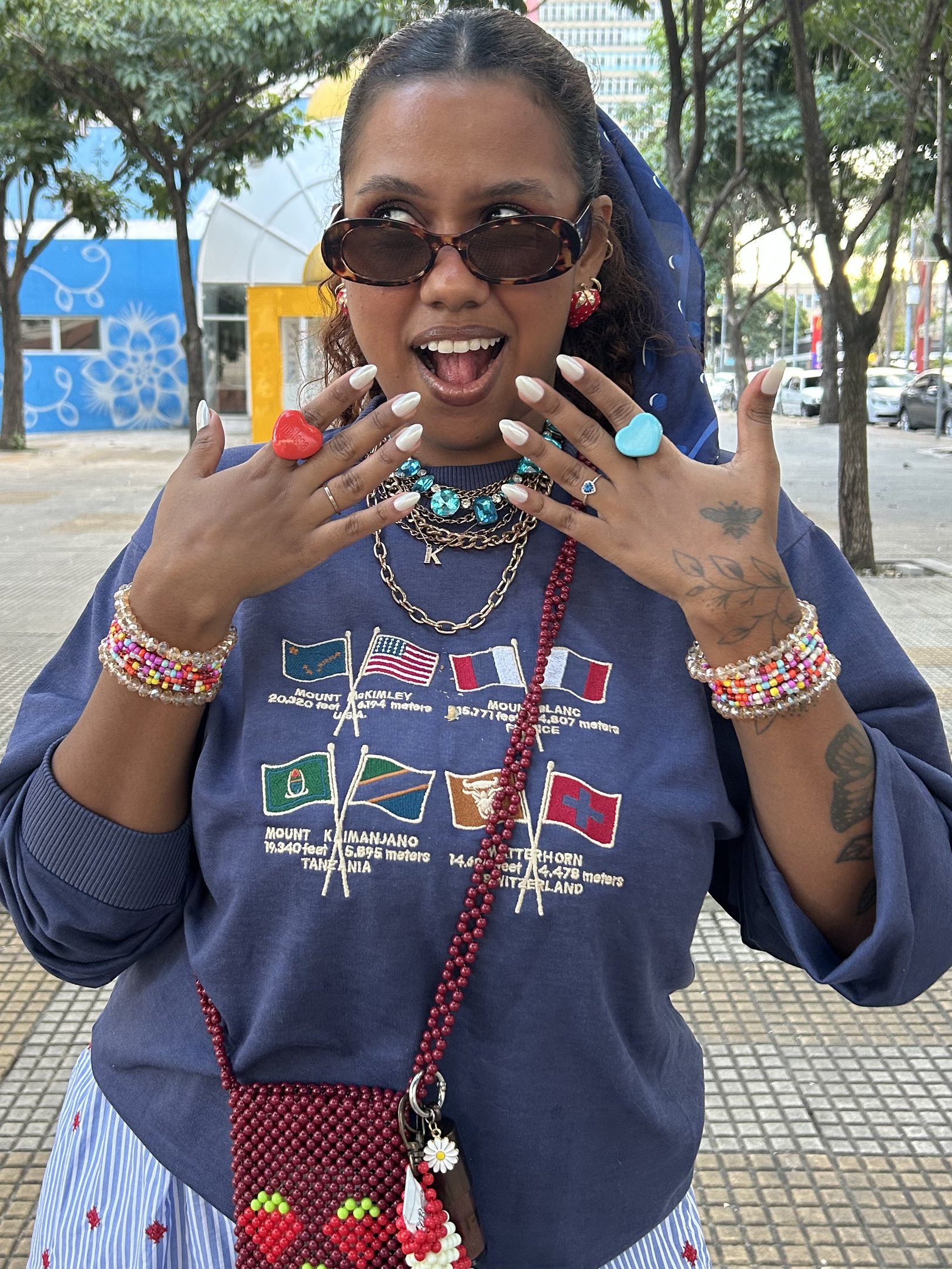 Mulher sorrindo exibe unha autocolante branca, usando anéis chamativos e pulseiras coloridas.
