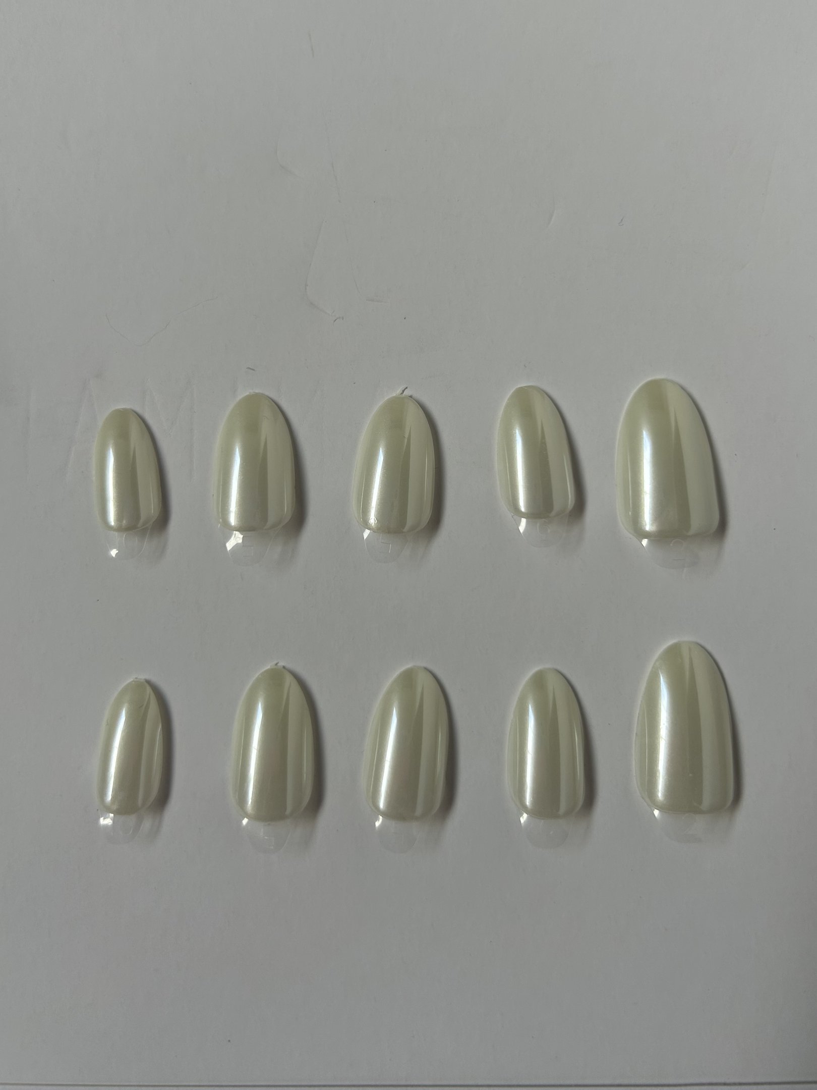 Nove unhas autocolantes em formatos ovais e tons claros dispostas sobre superfície branca.