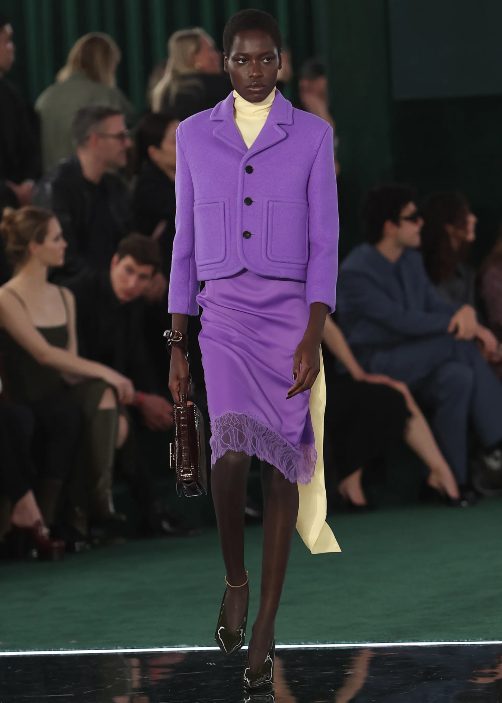 Modelo na passarela com traje lilás, provando que o roxo é tendência. Público ao fundo observando.