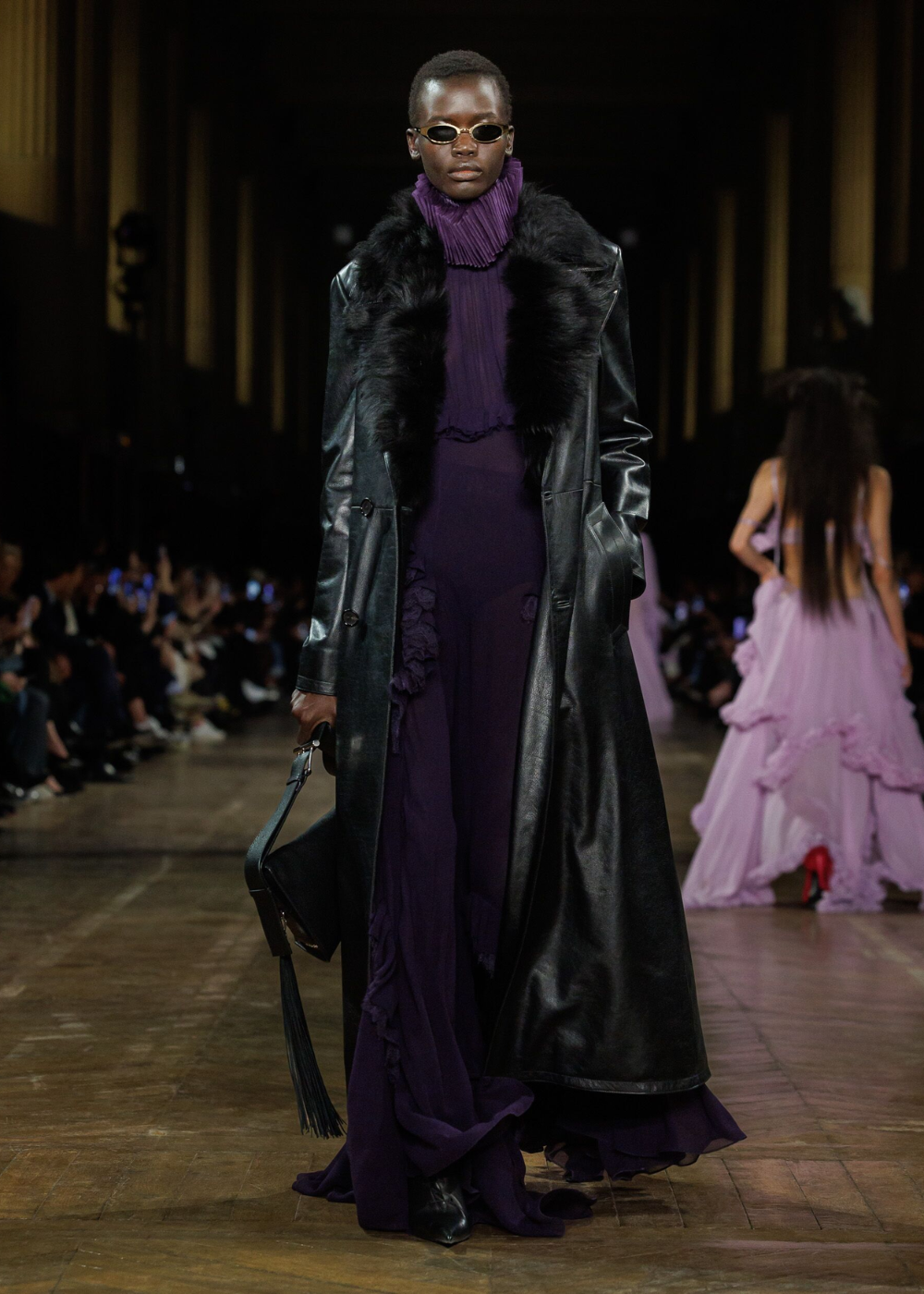 Modelo com casaco preto longo desfila com vestido roxo e bolsa. O roxo é tendência na passarela.