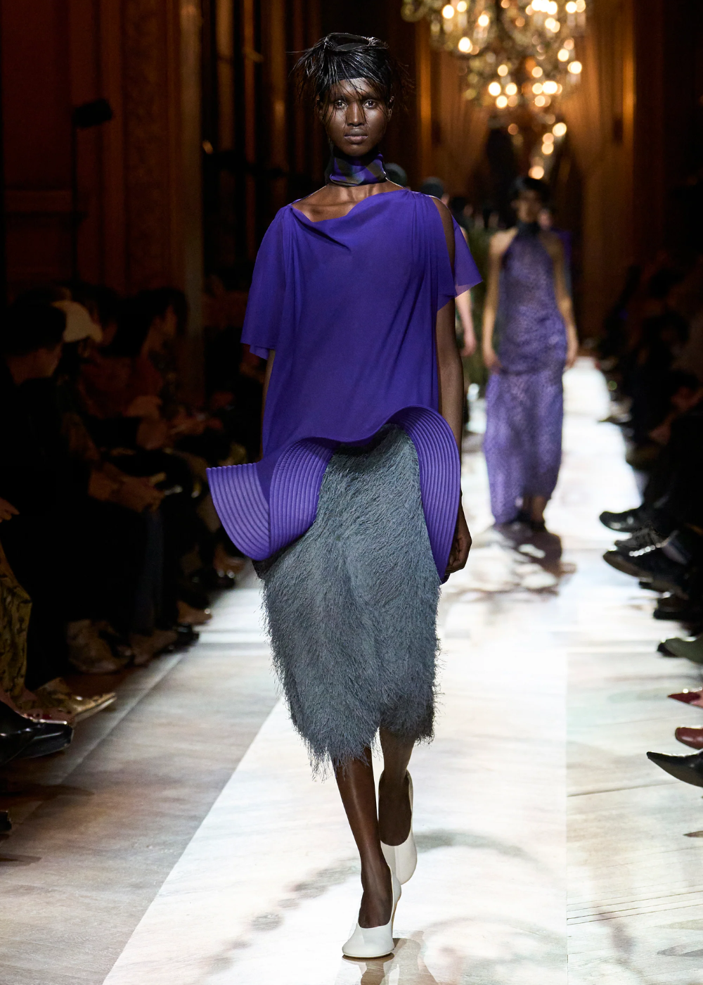 Modelo em traje com blusa roxa vibrante e saia texturizada cinza desfilando em passarela iluminada.