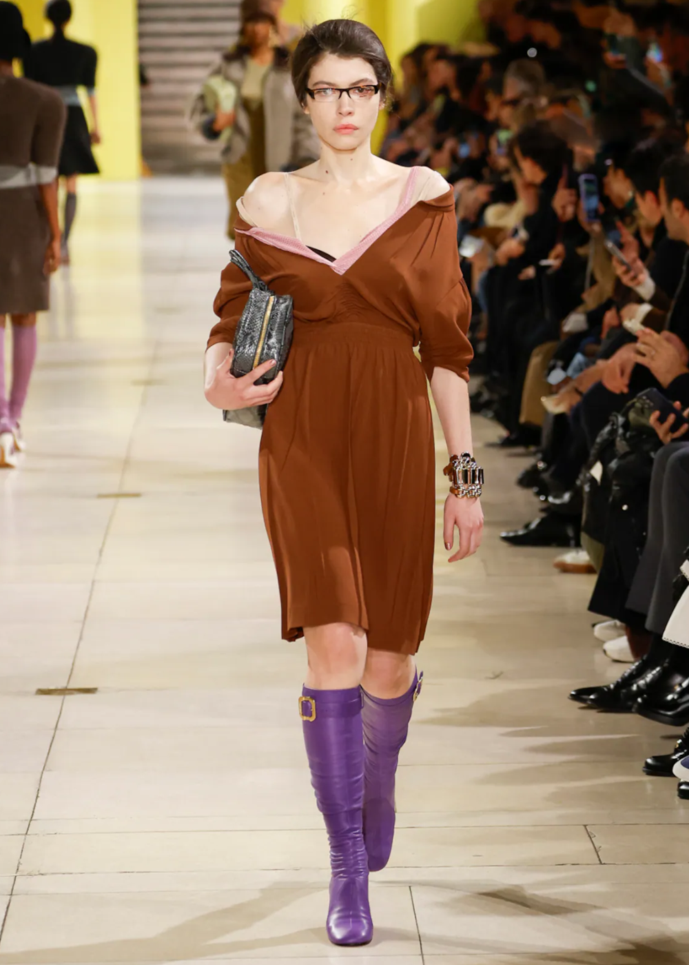 Modelo desfilando com vestido marrom e botas roxas em passarela, atraindo olhares atentos.
