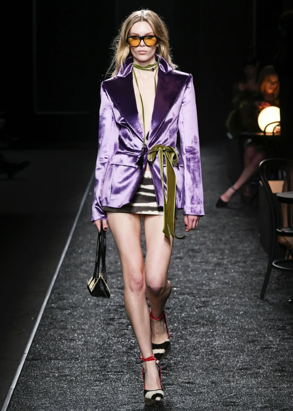 Modelo desfila em passarela usando blazer de veludo roxo brilhante e óculos de sol laranja.