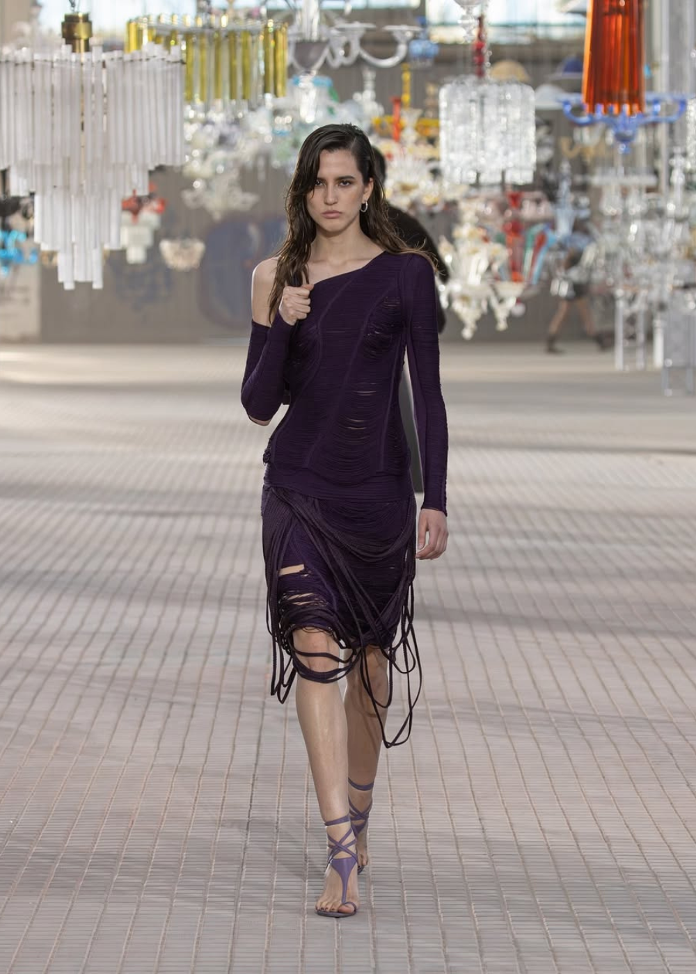 Modelo com vestido roxo texturizado em passarela, destacando tendência de moda com lustres ao fundo.