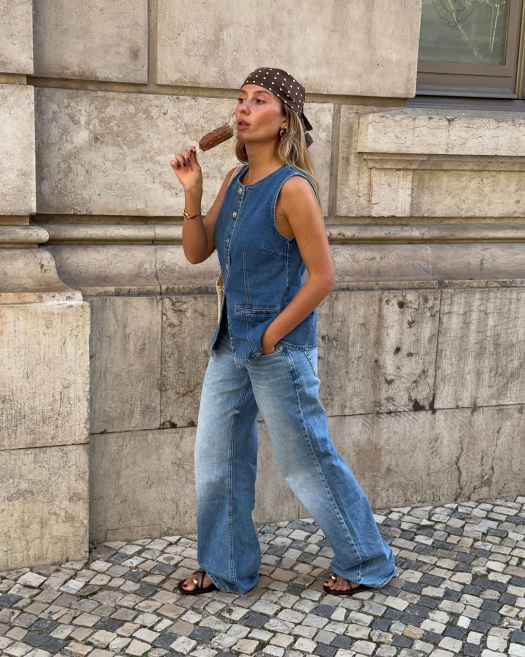 Mulher de lenço e jeans caminha, com sandália de dedo e picolé em mãos, em calçada de pedras.