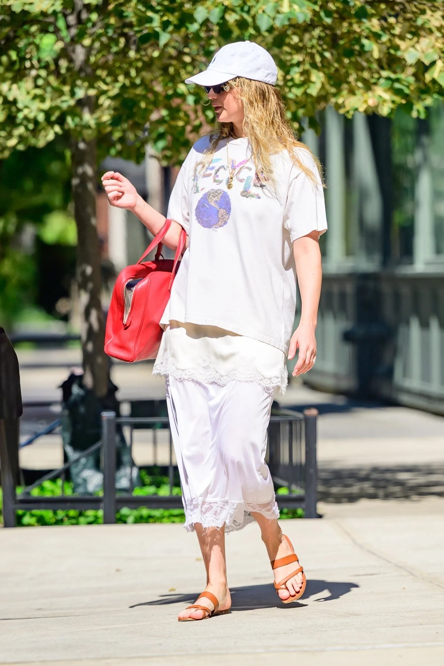 Pessoa caminhando na calçada, vestindo camiseta branca e sandália de dedo marrom, carregando bolsa vermelha.