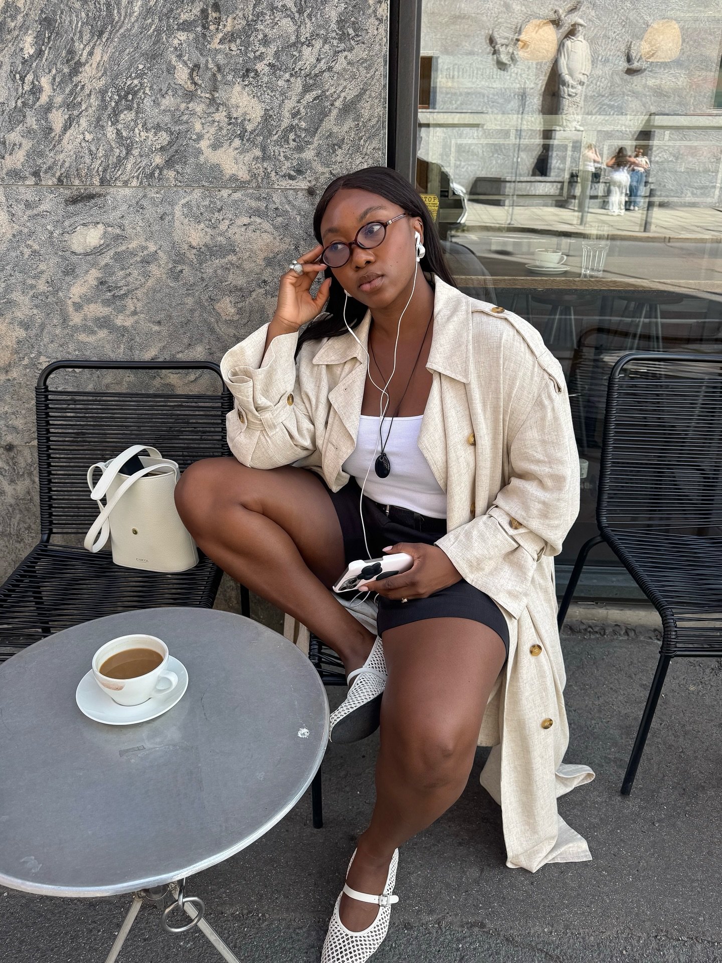 Mulher sentada em café ao ar livre, usando trench coat bege e sapatilha confortável branca.