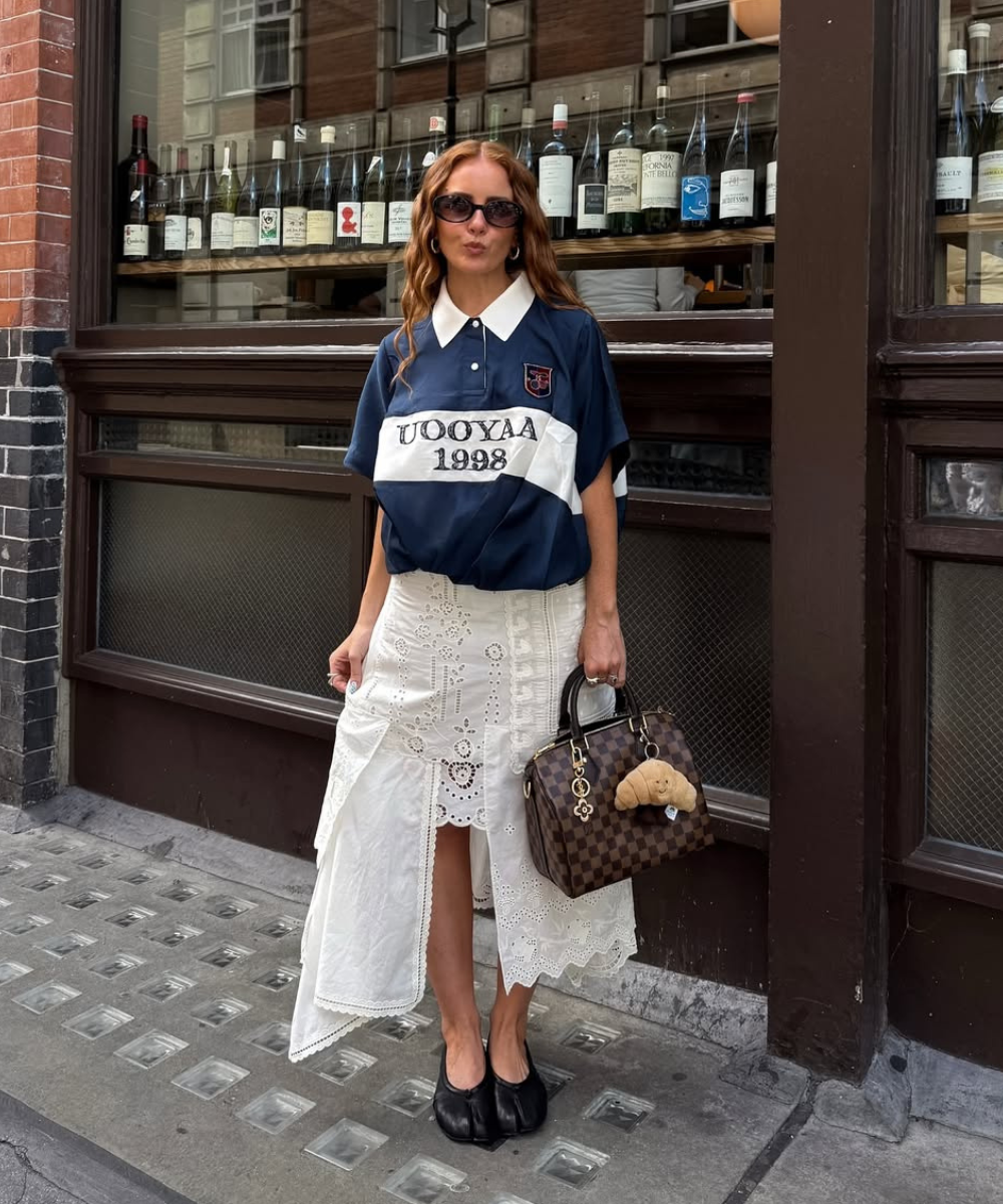 Mulher usa camisa azul e saia branca rendada, com bolsa marrom e sapatilhas estilosas. Fundo de garrafas expostas em vitrine.