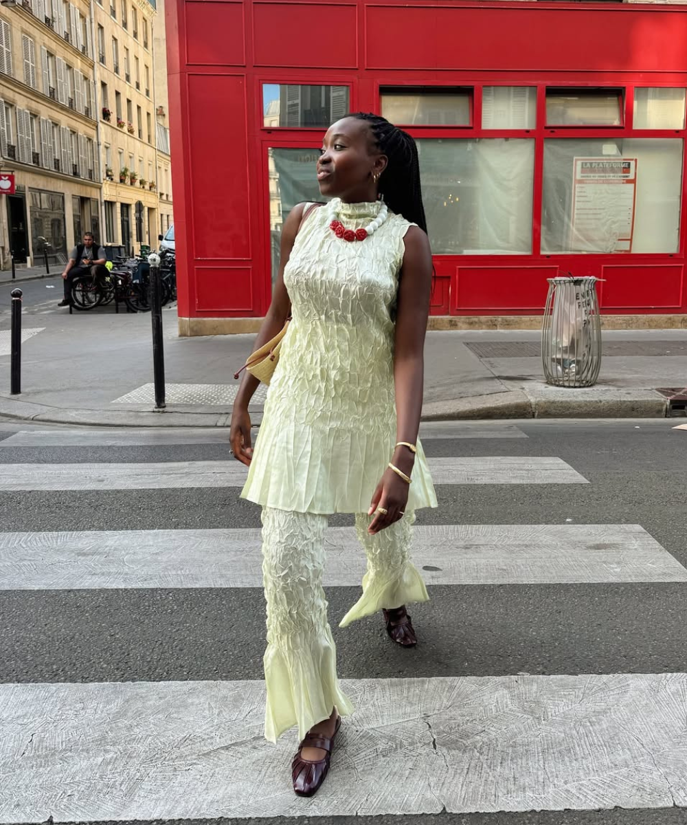 Mulher atravessando rua, vestida de verde com sapatilhas estilosas marrons e colar vermelho. Fundo urbano com parede vermelha.