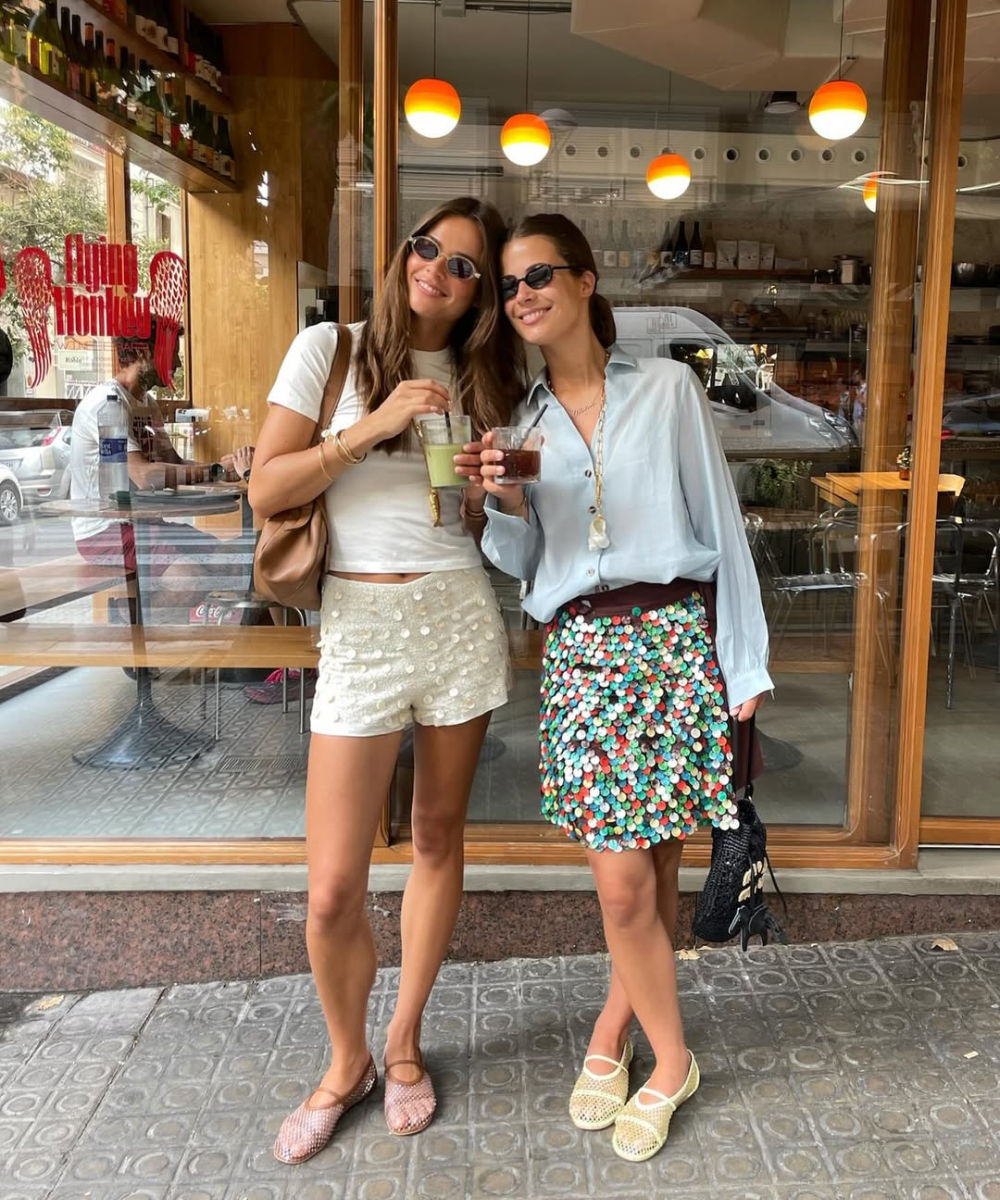 Duas mulheres sorridentes usam shorts e saias coloridos, segurando bebidas. Suas sapatilhas estilosas completam o visual descontraído.