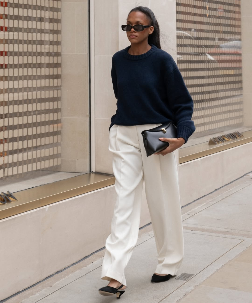 Pessoa com suéter azul, calça branca e sapato confortável e chique. Segura uma bolsa preta, caminhando em ambiente urbano.
