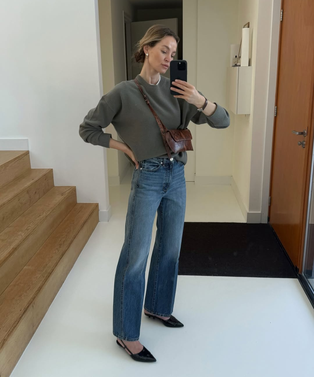 Pessoa vestindo jeans e suéter com sapato elegante, usando celular para tirar selfie em um ambiente interno minimalista.