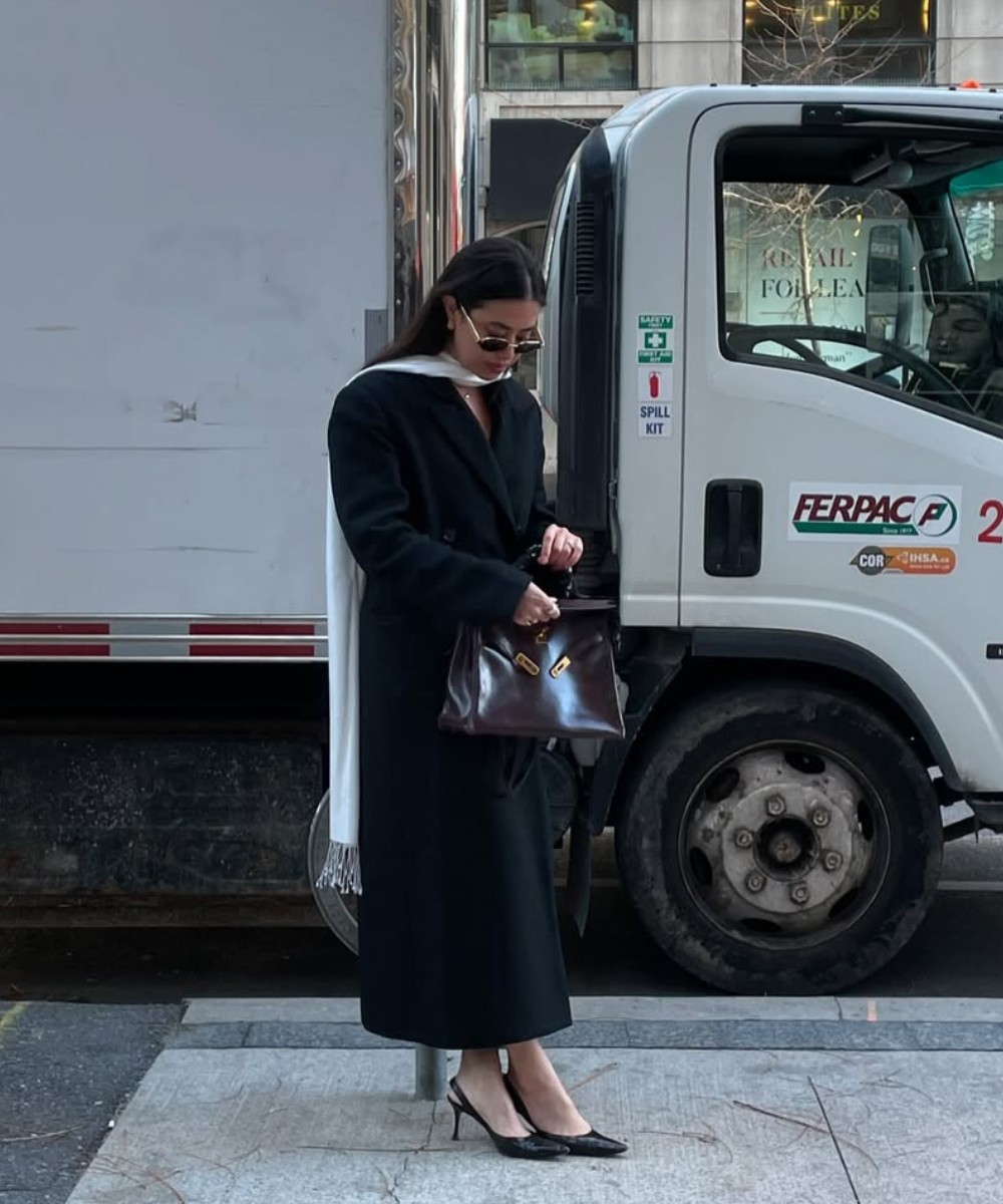 Mulher vestindo casaco preto e sapato elegante e chique, segurando bolsa marrom. Caminhão ao fundo.