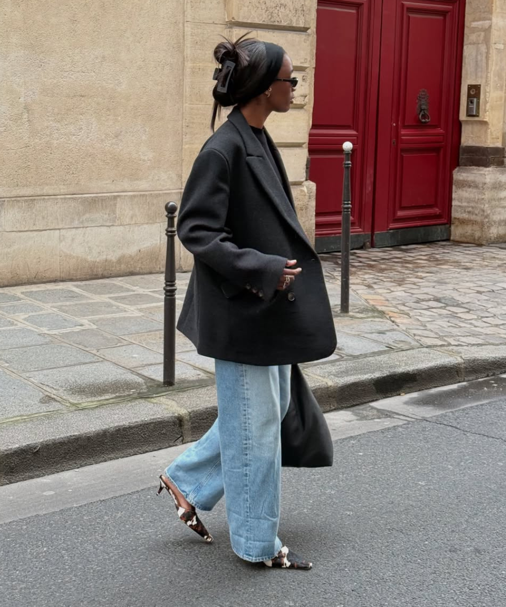 Pessoa com blazer preto, calça jeans larga e sapatos de salto com estampa cow print, caminhando em rua de paralelepípedos.