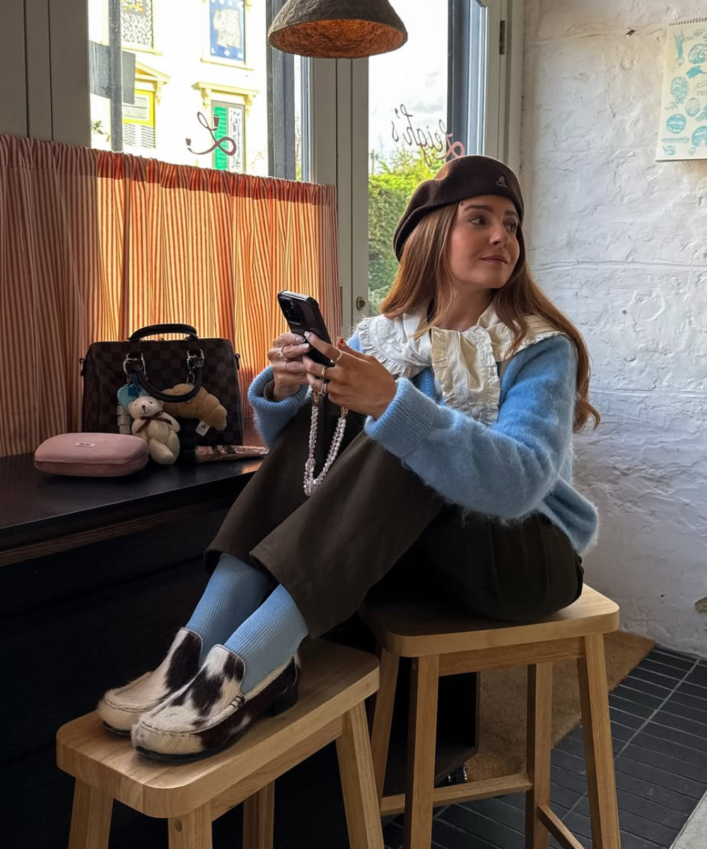 Mulher sentada em banqueta, segurando celular e usando sapatos com estampa cow print e  meias azuis.