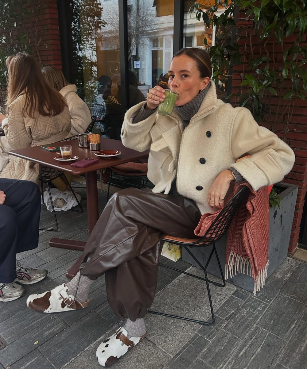 Pessoa com casaco bege e sapatos cow print, sentada em cafeteria ao ar livre, bebendo algo verde.