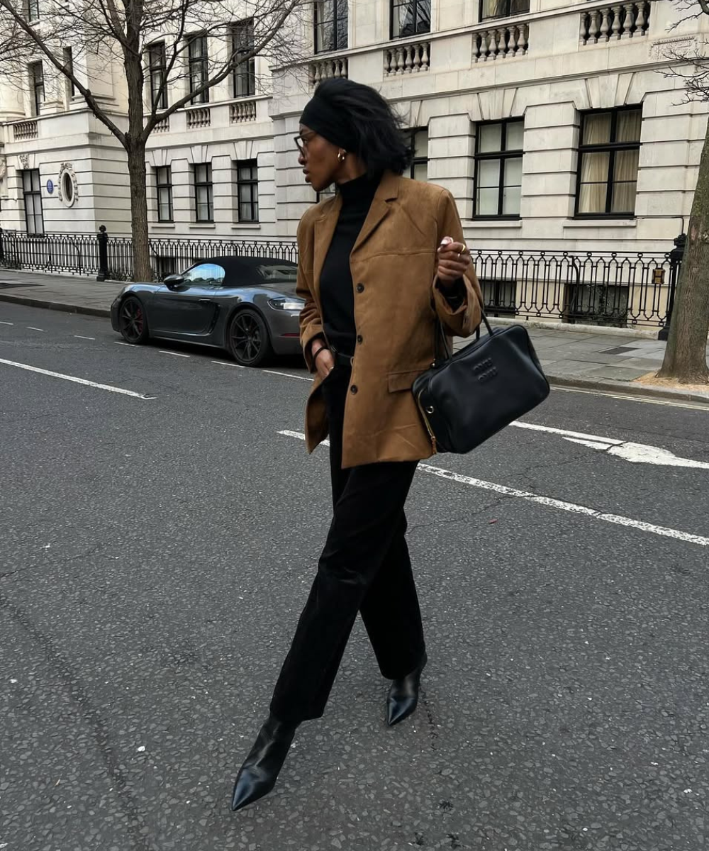 Pessoa com casaco marrom e sapatos de inverno elegantes caminha em rua urbana. Carro e edifício ao fundo.