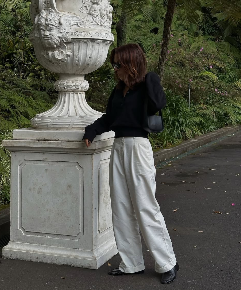 Pessoa com blusa preta e calça branca em frente a um vaso ornamental, vestindo sapatos elegantes pretos.