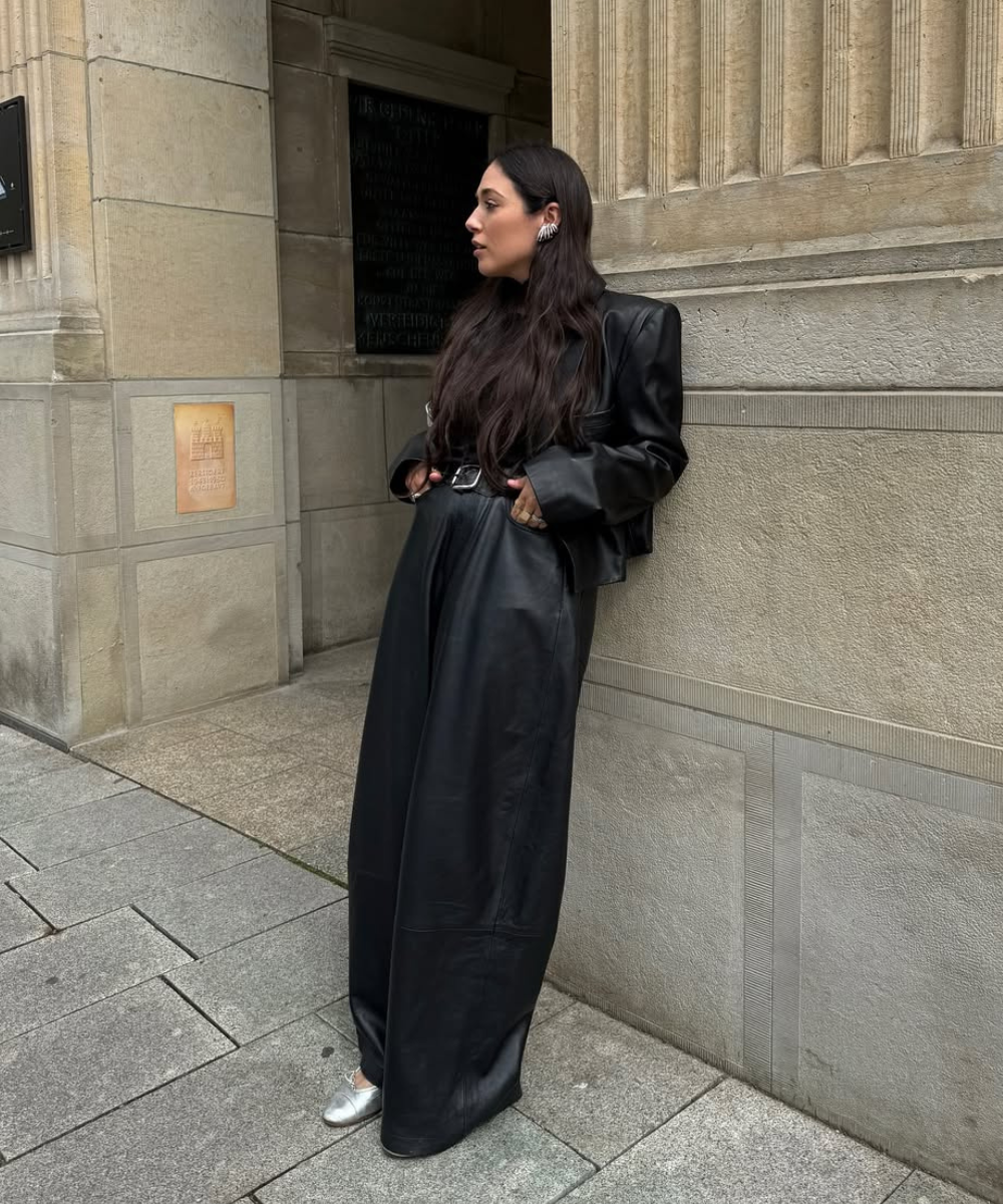 Pessoa usando conjunto de couro preto, encostada em parede de pedra. Sapatos prateados completam o visual.