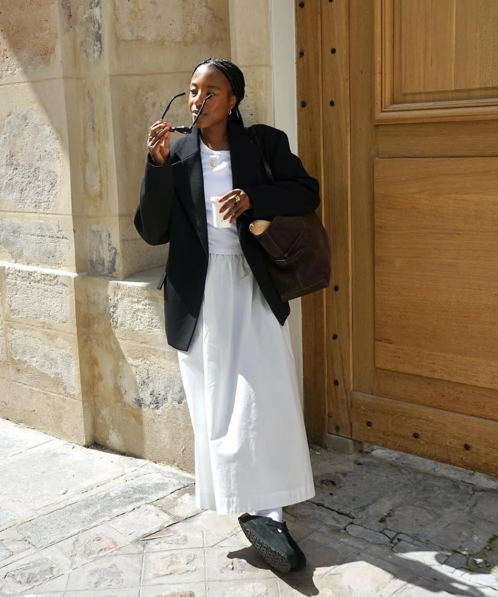 Pessoa com óculos de sol, casaco preto e saia longa branca. Sapatos de inverno elegantes completam o visual descontraído.