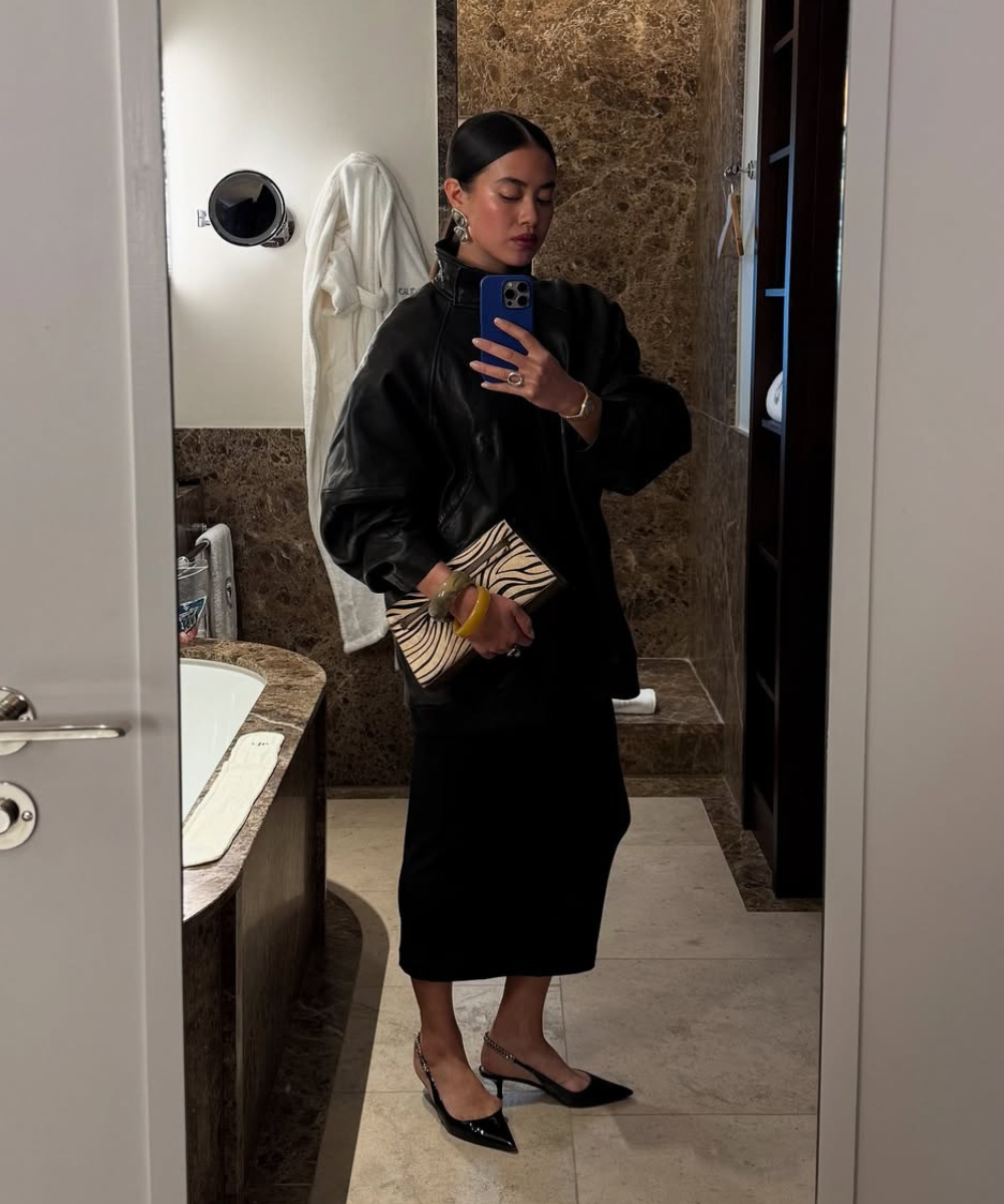 Usuário com vestimenta preta elegante em banheiro, segurando clutch listrada. Sapatos de salto pontudo.