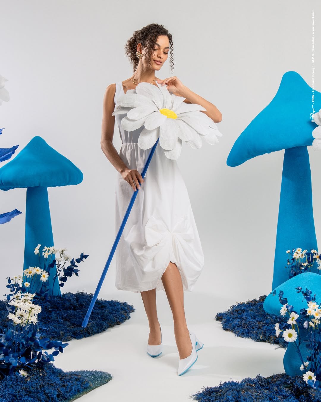 Mulher com vestido branco segura flor gigante, cercada por cogumelos azuis e flores. Usa sapato branco.