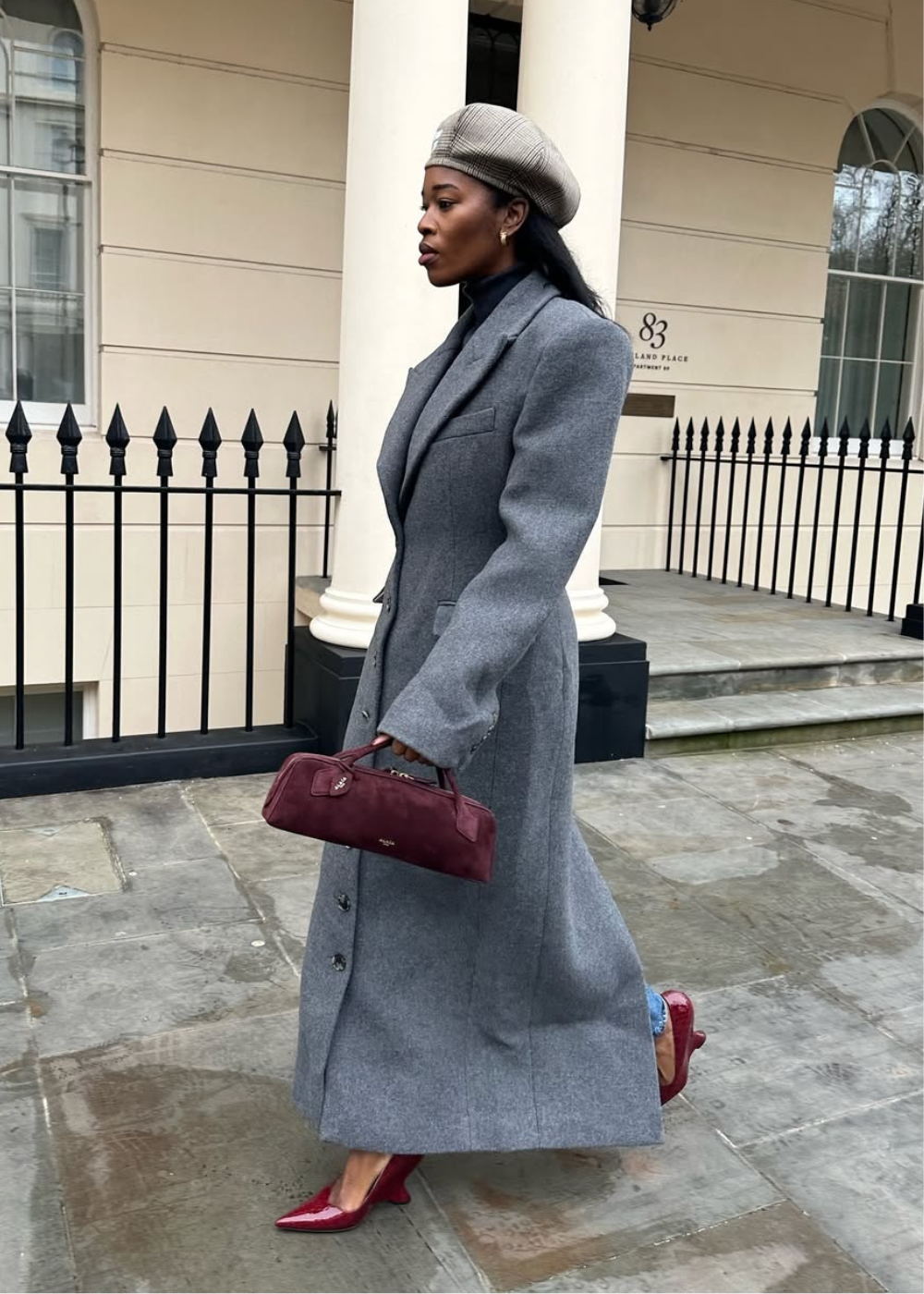 Mulher com casaco cinza e boina bege caminha usando scarpin anabela vermelho, segurando bolsa vinho.