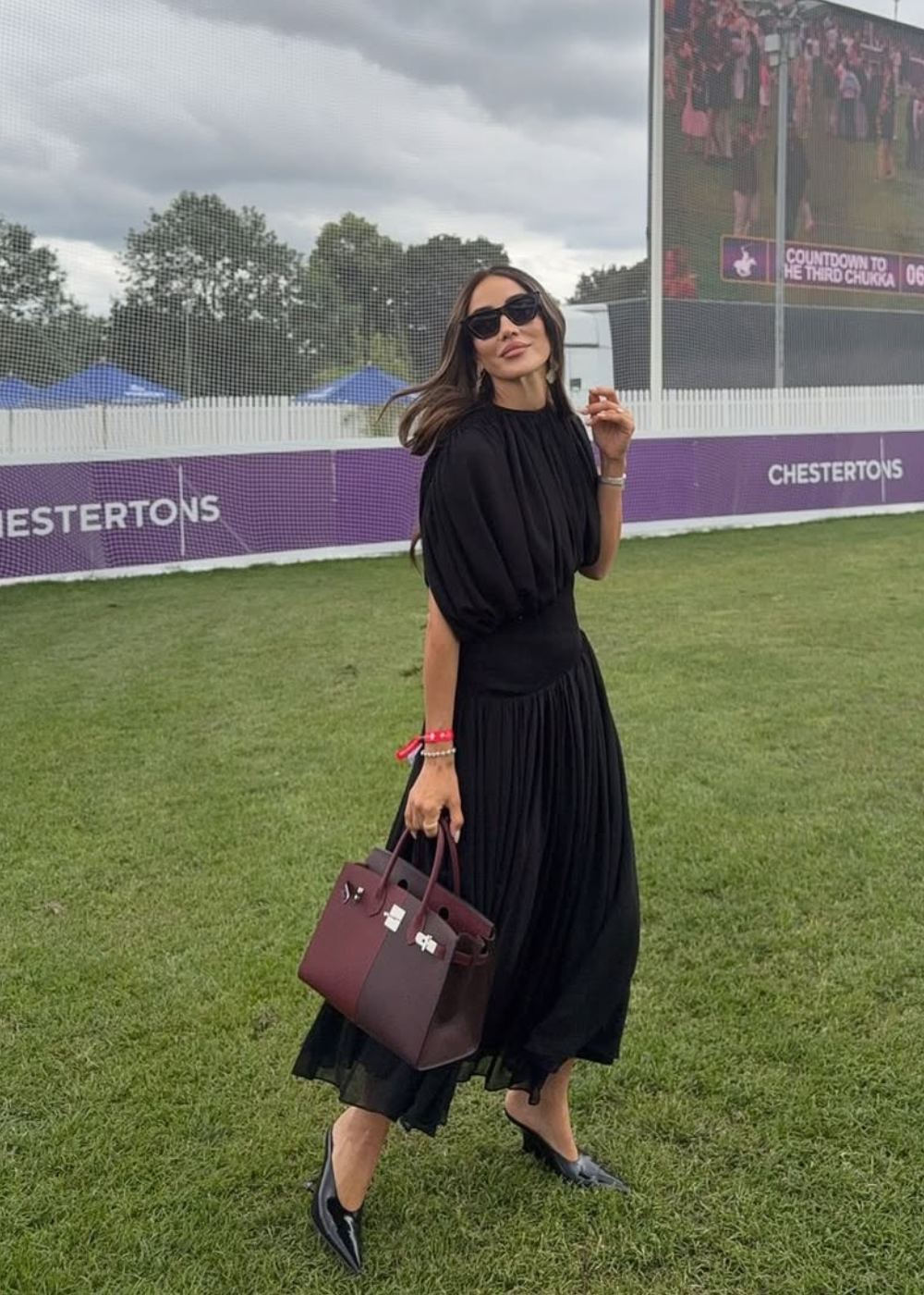 Mulher elegante com vestido preto e bolsa vinho caminha em evento ao ar livre, utilizando scarpin de salto moderado.