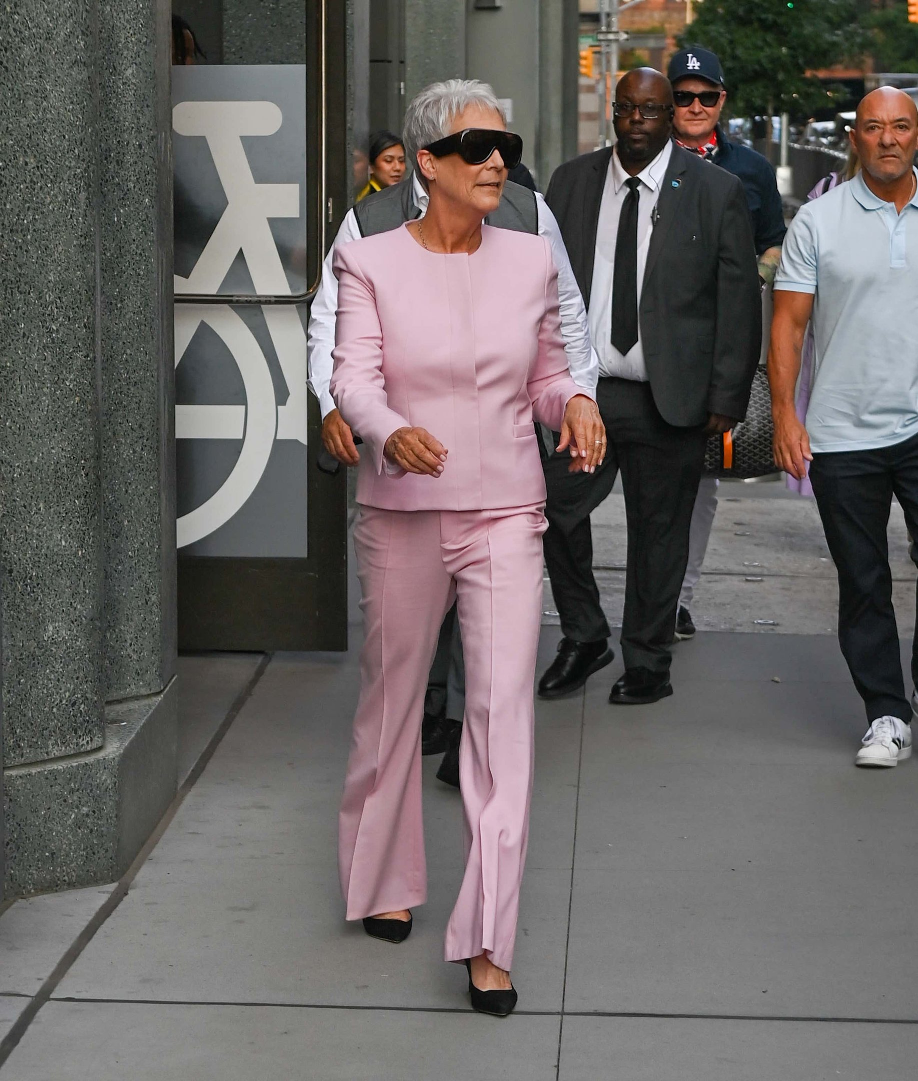 Pessoa de terno rosa caminha acompanhada por outras, em ambiente urbano. Sexta-feira muito louca 2