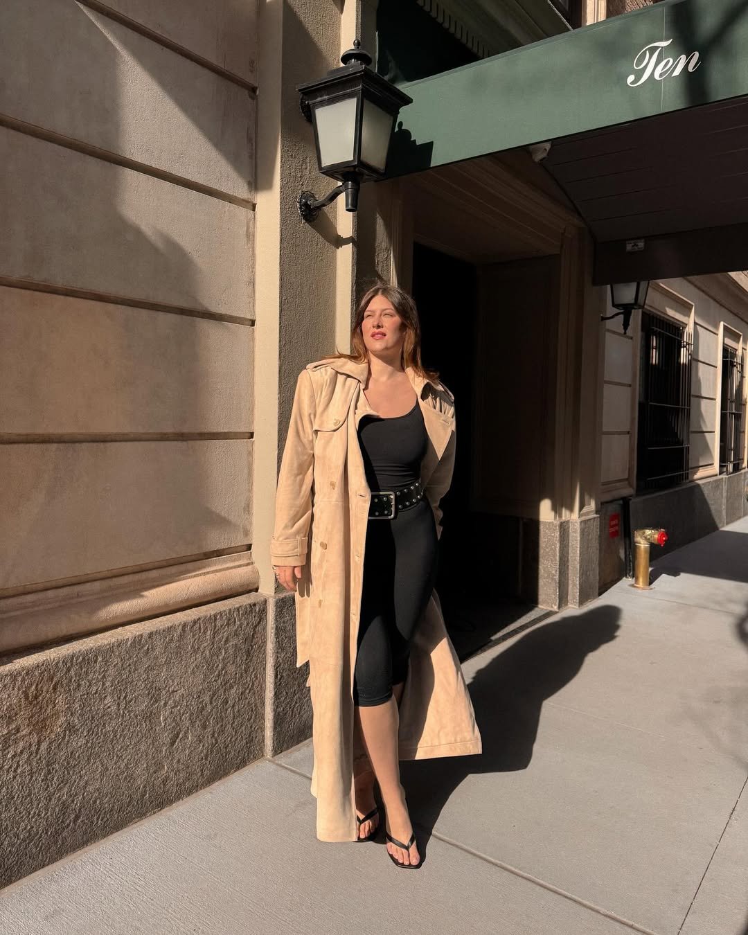 Mulher em um look sexy e elegante com casaco bege, roupa preta e sandálias sob luz solar, em uma calçada urbana.