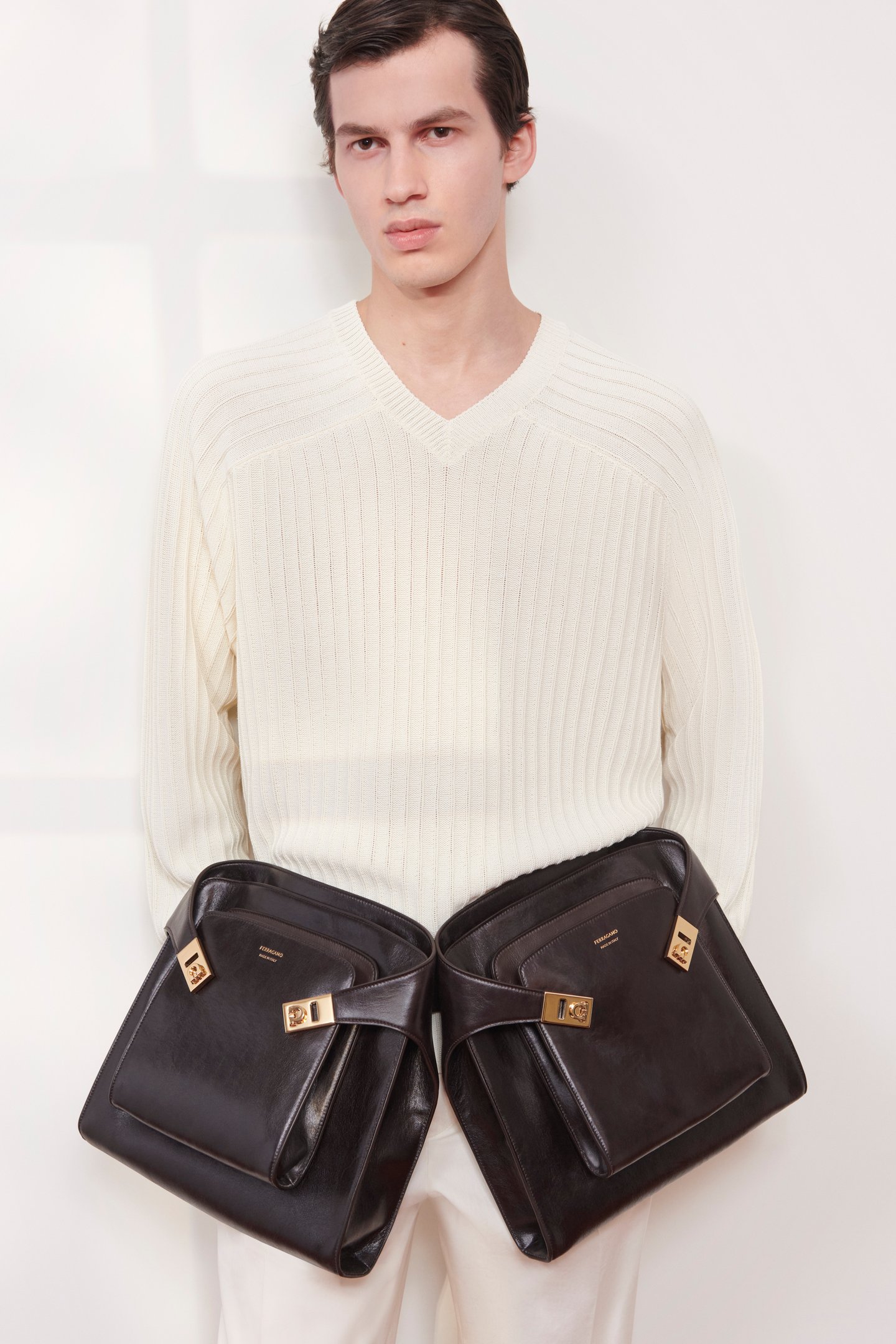 Modelo com suéter creme segura duas bolsas Ferragamo de couro marrom com detalhes em dourado.