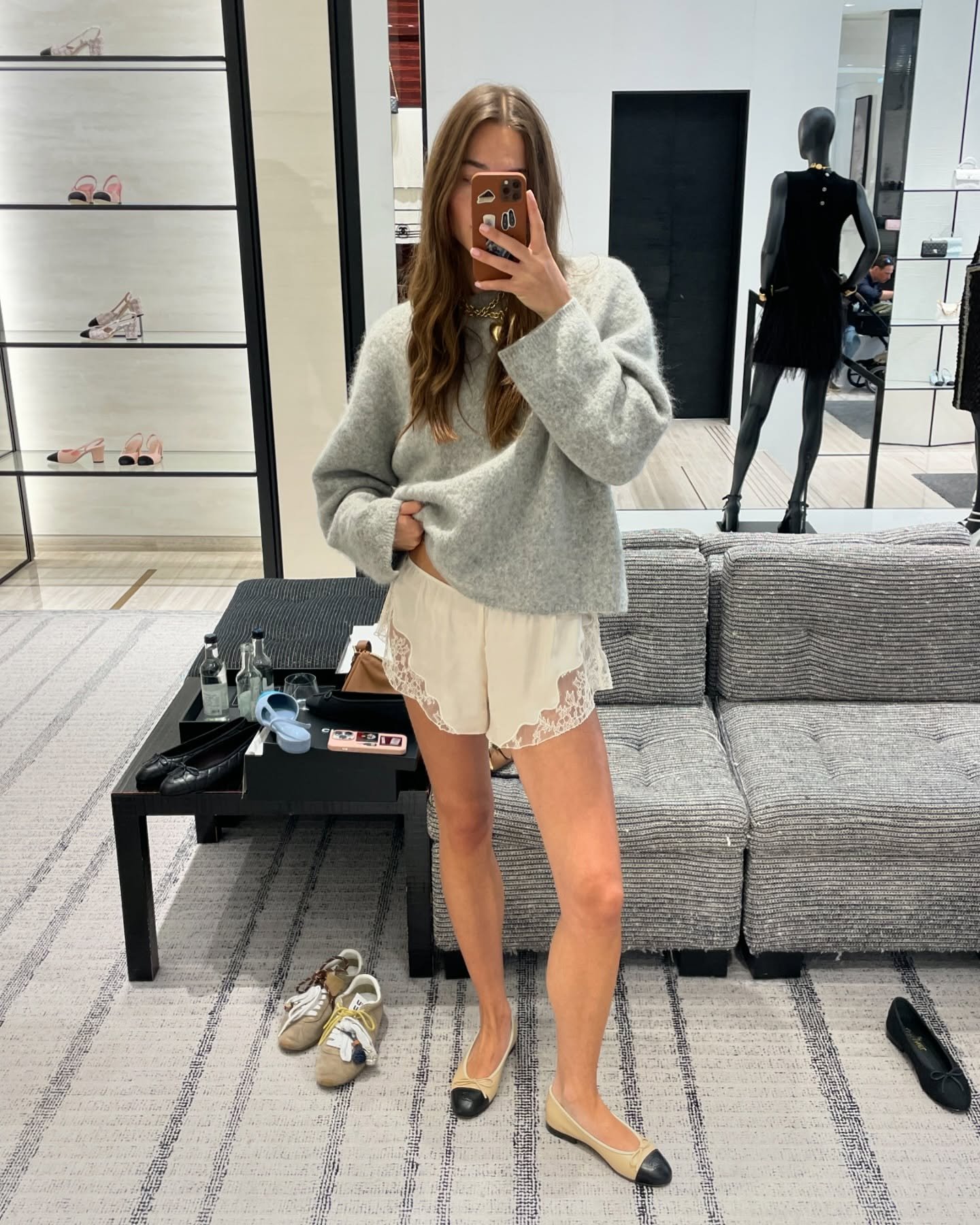 Pessoa em provador, vestindo blusa cinza e short de pijama com renda, tirando selfie no espelho.