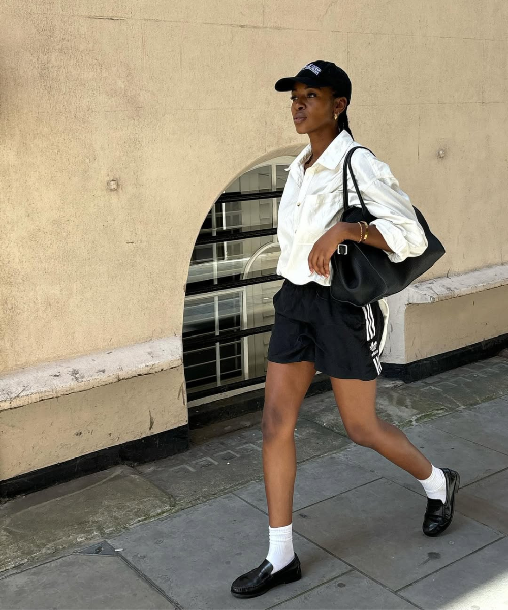Pessoa caminhando na calçada com camisa branca e shorts preto em alta, completando o look com bolsa e boné.