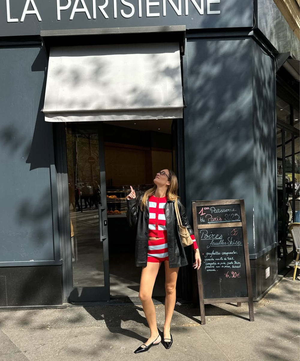 Mulher em frente a padaria, usando blazer preto e shorts tendência listrados vermelho e branco, apontando para cima.